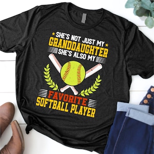 以下が含まれることがあります： ソフトボールのグラフィックがプリントされた濃いグレーのTシャツ。デザインには、黄色のソフトボール、交差したバット、そして「彼女は私の孫娘だけでなく、私のお気に入りのソフトボール選手でもあります」というテキストが含まれています。