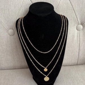 May include: A gold-colored, three-strand necklace displayed on a black mannequin. The necklace features delicate chains with two circular pendants, one smooth and one textured. The background is a light gray fabric.