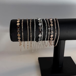 May include: A display of various delicate gold and silver chain bracelets. The bracelets are of different styles, including disc, link, and beaded designs. They are arranged on a black jewelry display stand against a white background.