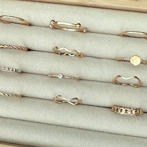 May include: A selection of gold-coloured rings in diverse styles, such as a twisted band, a band with small spheres, and a band with a small clear stone. The rings are presented in a light grey jewellery box.