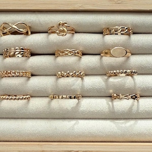 May include: A collection of gold-colored rings displayed in a jewelry box. The rings feature various designs, including chain links, knots, and geometric shapes. The rings are arranged on a light gray, velvety surface.