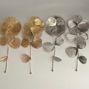 May include: Four metal flower wall art pieces in gold, bronze, and silver tones. Each flower has four petals and a stem with leaves. The flowers are made of thin wire and have a delicate, airy appearance.