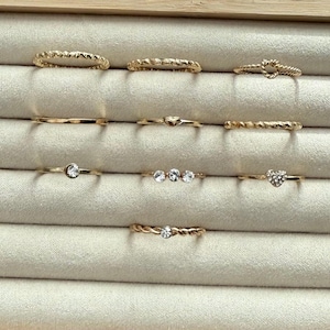 May include: A collection of gold-toned rings displayed in a jewelry box. The rings feature various designs, including textured bands, heart shapes, and small gemstones. The rings are arranged on a light beige, velvety surface.