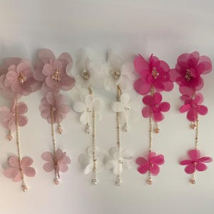 May include: A collection of floral hair accessories in shades of pink and white. Each piece features layered fabric flowers, embellished with beads and dangling chains. The accessories are designed for special occasions and add a touch of elegance.