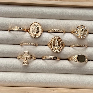 May include: A collection of gold-colored rings displayed in a jewelry box. The rings feature various designs, including religious symbols like crosses and depictions of the Virgin Mary. Some rings have gemstone accents.