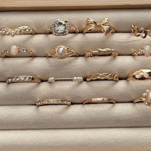May include: A collection of gold-toned rings displayed in a jewelry box. The rings feature various designs, including gemstones, pearls, and intricate patterns. The rings are arranged on a beige fabric.