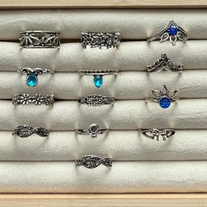 May include: A collection of silver-toned rings displayed on a cream-colored surface. The rings feature various designs, including floral, crown, and infinity symbols, and some are adorned with blue gemstones. The rings are arranged in rows.