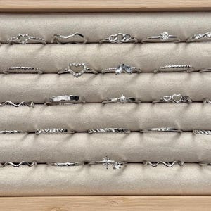 May include: A collection of silver rings displayed in a wooden jewelry box. The rings feature various designs, including wave patterns, hearts, and gemstones. The rings are arranged on a light beige, textured surface.