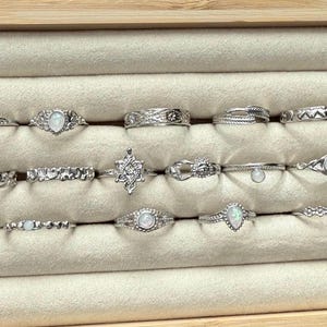 May include: A collection of silver rings with various designs, including bands with intricate patterns, opalescent stones, and delicate shapes. The rings are displayed in a beige jewelry tray.