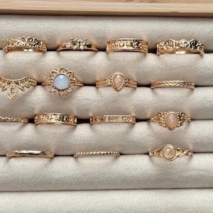 May include: A collection of gold-colored rings displayed in a jewelry box. The rings feature various designs, including floral, geometric, and gemstone accents. The rings are arranged on a light gray, velvet-lined surface.
