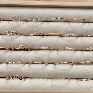 May include: A collection of gold-colored rings displayed in a jewelry box. The rings feature various designs, including twisted bands, heart shapes, and small gemstones. The rings are arranged on a light beige, velvet-like surface.
