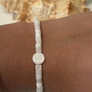 May include: White beaded bracelet with a circular centerpiece. The bracelet is made of small, rectangular beads and a round, flat centerpiece. The bracelet is worn on a wrist, with a seashell in the background.