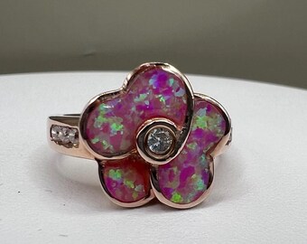 May include: A rose gold flower ring with a pink opal center and a small diamond in the middle. The ring has a band with small diamonds. The flower petals have green and purple accents.