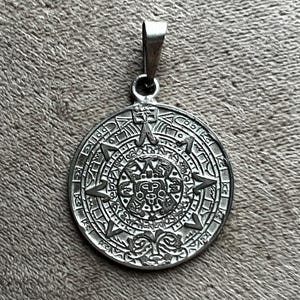 May include: A silver-colored pendant with an intricate Aztec calendar design. The circular pendant features detailed carvings of symbols and figures, with a loop at the top for a chain. The pendant is displayed against a textured, neutral background.
