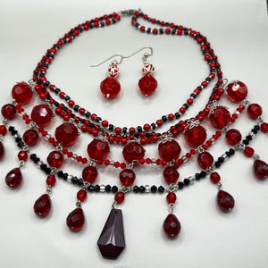 May include: A red and black beaded necklace and earring set. The necklace features multiple strands of red and black beads, with larger red faceted beads and teardrop pendants. The earrings have red faceted beads.
