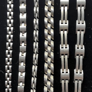 May include: A collection of silver-toned metal bracelets with various link designs. The bracelets are displayed against a black background, showcasing their different styles, including chain and rectangular link patterns. The image highlights the jewelry's metallic sheen.
