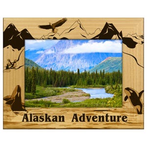 May include: Wooden picture frame with an Alaskan landscape scene. The frame features etched images of mountains, an eagle, and orcas. The text "Alaskan Adventure" is at the bottom of the frame.
