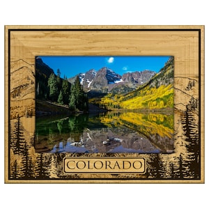 May include: A wooden picture frame with a scenic landscape of mountains, a lake, and trees. The frame has a light brown wood grain finish with etched mountain and tree designs. The word "COLORADO" is displayed on the bottom.