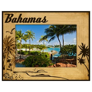 May include: Wooden picture frame with a scenic view of the Bahamas. The frame has the word "Bahamas" in a cursive font at the top. The image inside shows palm trees, blue water, and a bright sky. Decorative palm tree silhouettes are on the frame.