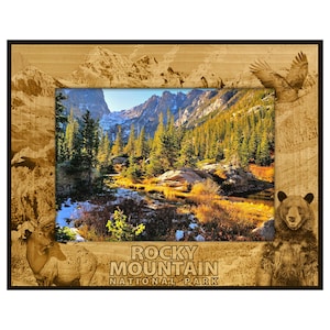 May include: Wooden framed art print featuring a scenic view of the Rocky Mountain National Park. The frame has etched mountain and wildlife designs, including an elk, bear, and eagle. The print displays a mountain landscape with trees and a river, and the text "Rocky Mountain National Park".
