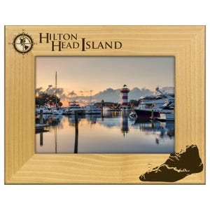 May include: A light-colored wooden picture frame with the words "Hilton Head Island" and a compass rose. The frame holds a scenic photo of a harbor with boats, a lighthouse, and a sunset.