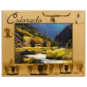 May include: Wooden picture frame with a scenic landscape of a river and mountains. The frame has the word "Colorado" in script, along with decorative elements like a longhorn skull, a teepee, and a cowboy. Silhouettes of cattle are at the bottom.
