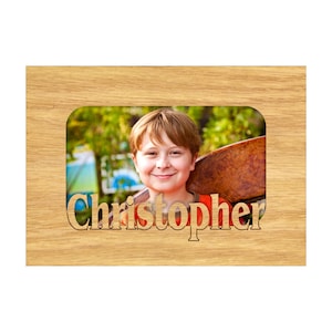 May include: Wooden picture frame with a photo of a smiling child. The frame is light brown with the name "Christopher" cut out at the bottom. The photo shows a boy with red hair and an orange shirt.