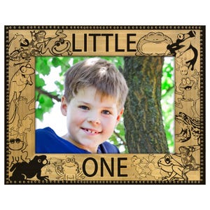 May include: Wooden picture frame with the words "LITTLE ONE" and frog illustrations. The frame has a light brown wood grain finish with black frog designs and a black border. The frame holds a photo of a smiling child.