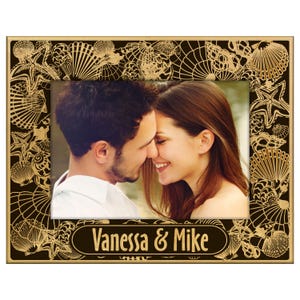 May include: A rectangular photo frame with a black background and a gold-colored seashell and starfish design. The frame holds a photo of a couple. The text "Vanessa & Mike" is displayed at the bottom of the frame.