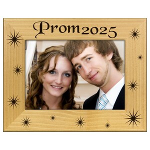 May include: A light-colored wooden picture frame with the text "Prom 2025" in a decorative font. The frame has a photo of a smiling couple and starburst designs.