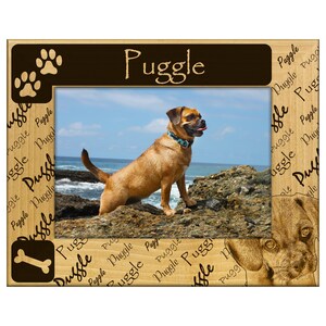 May include: Wooden photo frame featuring a Puggle dog. The frame has the word "Puggle" in a brown banner at the top. The frame also has paw prints and a dog bone icon. The frame has a picture of a Puggle dog on a rocky beach.