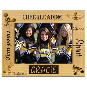 May include: A wooden picture frame with the word "CHEERLEADING" at the top. The frame surrounds a photo of three smiling cheerleaders in black and yellow uniforms. The frame has the words "Pom poms", "Basket toss", "Cheer", and "Spirit" engraved on it.