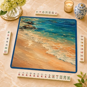 May include: A square mahjong mat featuring an impressionistic oil painting of a sandy beach with turquoise ocean waves and dark rocks. The mat is surrounded by four rows of white mahjong tiles with red, green, and black characters and symbols.
