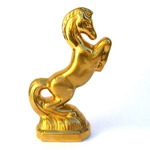 Puede incluir: Una figura dorada de caballo en posición encabritada. La escultura está hecha de un material brillante y reflectante, y está montada sobre una base rectangular. La melena y la cola del caballo están detalladas, y el diseño general es elegante.