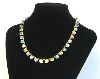 May include: A gold-toned necklace with iridescent, round crystals. The crystals are evenly spaced along the chain, creating a sparkling, elegant look. The necklace is displayed on a black mannequin.
