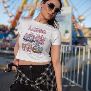 May include: White t-shirt featuring the word "BABUBU" in pink, above a cartoon graphic of four sleeping characters in pink and blue boxes. The person is wearing black shorts and a black and white checked shirt.