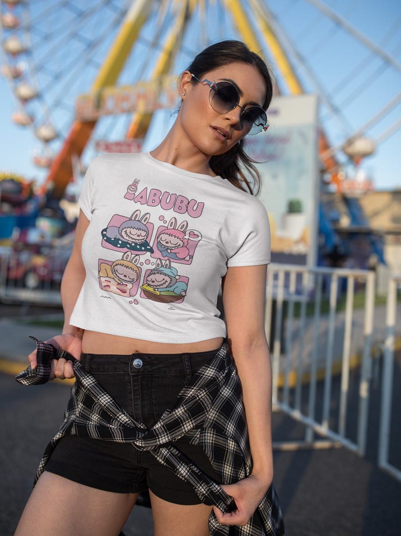 May include: White t-shirt featuring the word "BABUBU" in pink, above a cartoon graphic of four sleeping characters in pink and blue boxes. The person is wearing black shorts and a black and white checked shirt.