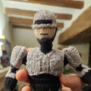 May include: Hand-knitted Robocop doll in gray, black, and tan. The doll features a helmet, body armor, and articulated limbs, all crafted from yarn. The figure is held in a person's hand, showcasing the detail of the handmade item.