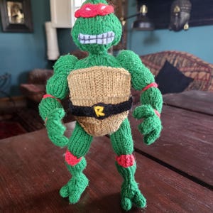 Op de afbeelding: Handgebreide groene en bruine schildpadfiguur met een rode hoofdband, zwarte riem met een gele "R" en rode accenten. De figuur heeft een lachend gezicht met witte tanden. Een uniek, handgemaakt item.
