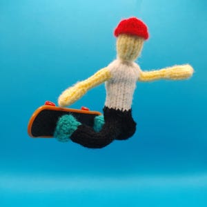 May include: A handmade knitted figure of a person skateboarding. The figure is wearing a red hat, white shirt, black pants, and teal shoes. The skateboard is black with orange and red accents. The background is a solid blue.