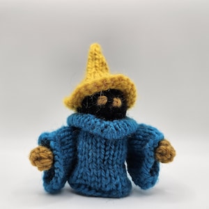 Puede incluir: Muñeco de mago tejido a mano. El muñeco tiene una cara negra con dos agujeros para los ojos, un sombrero puntiagudo amarillo y una túnica azul con detalles amarillos. El muñeco está hecho de hilo.