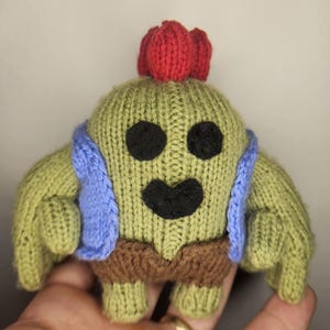 May include: A knitted cactus character in green, brown, blue, and red. The plush toy has a smiling face with black eyes and mouth, and a red headpiece. The character is wearing a blue vest and brown shorts.