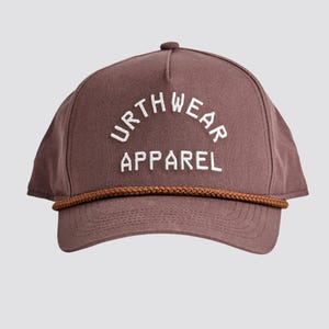 May include: A brown baseball cap with the text "URTHWEAR APPAREL" in white. The cap has a brown rope detail along the brim and a curved bill. The hat is a solid color.