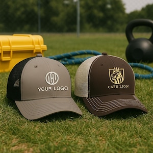 May include: Two baseball caps on green grass. One is grey with a black mesh back and the text "YOUR LOGO". The other is brown with a beige mesh back and a gold lion logo with the text "CAFE LION".