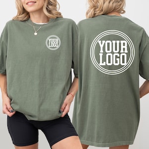 May include: Olive green t-shirts with the text "YOUR LOGO" printed in white on the front and back. The shirts are oversized and have short sleeves. The model is wearing black shorts.