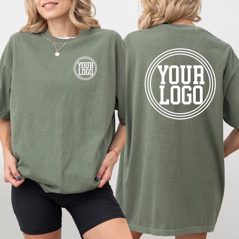 May include: Olive green t-shirts with the text "YOUR LOGO" printed in white on the front and back. The shirts are oversized and have short sleeves. The model is wearing black shorts.