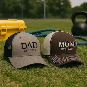 May include: Two baseball caps with mesh backs. One is gray with "DAD EST. 2025" in black text. The other is brown with "MOM EST. 2025" in white text. Both caps are on a grassy surface.