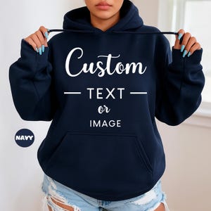 Peut inclure: Sweat à capuche bleu marine avec une poche avant et un cordon de serrage. Le texte "Custom TEXT or IMAGE" est imprimé en blanc. Un petit cercle bleu marine avec le mot "NAVY" est dans le coin inférieur gauche.