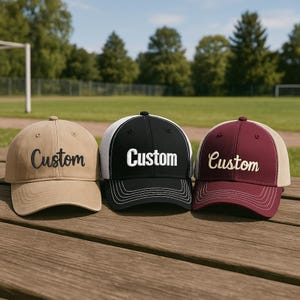 May include: Three baseball caps with the word "Custom" embroidered on the front. One cap is tan, one is black with a white mesh back, and the third is maroon with a tan mesh back. The caps are displayed on a wooden surface.