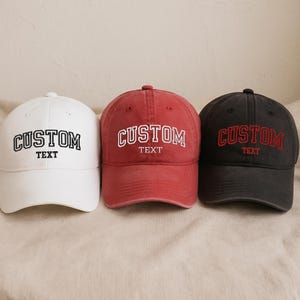 May include: Three baseball caps in white, red, and black. Each cap has the word "CUSTOM" embroidered in a contrasting color, with "TEXT" below. The caps are arranged on a neutral-toned surface.